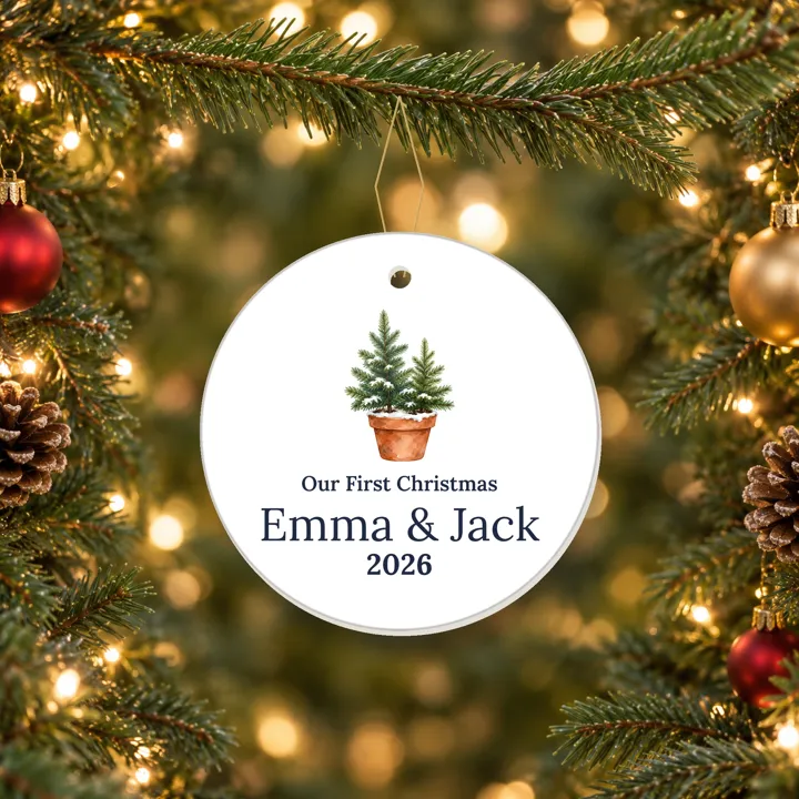 Ceramic Our First Christmas 2026 ornament with two potted evergreens and sample couple names, hanging on a tree